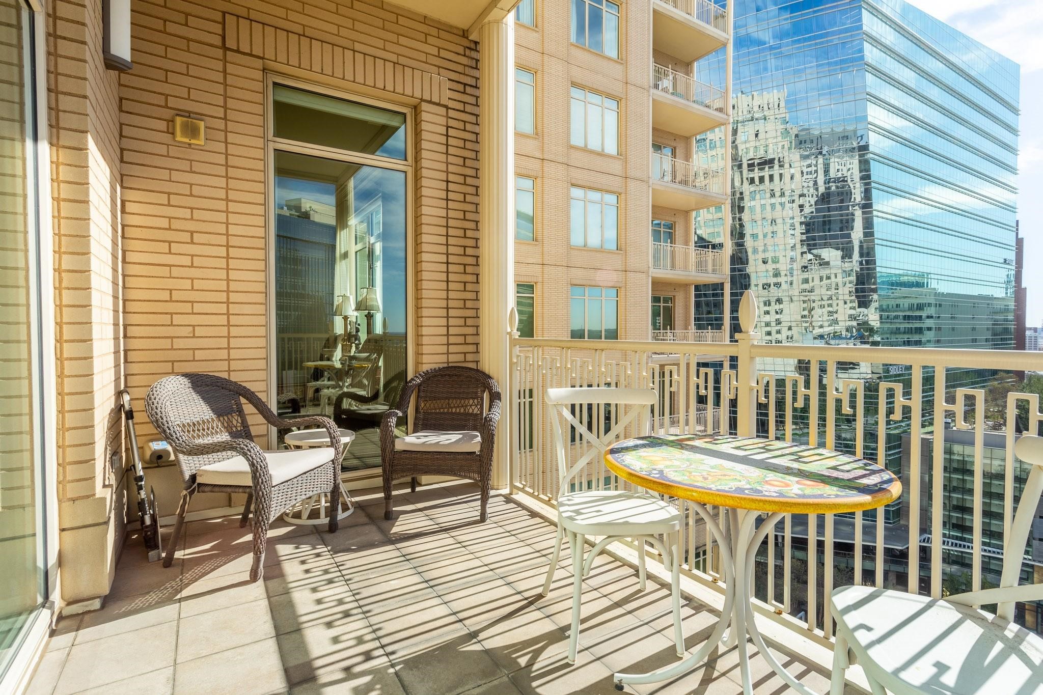 Furnished high-rise balcony with wicker chairs and a colorful table overlooking downtown Dallas towers.