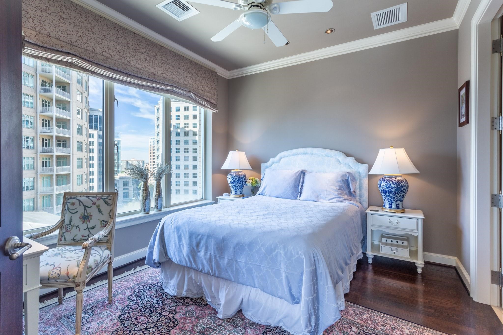 Furnished bedroom with a large bed, patterned armchair, and floor-to-ceiling windows overlooking downtown buildings.