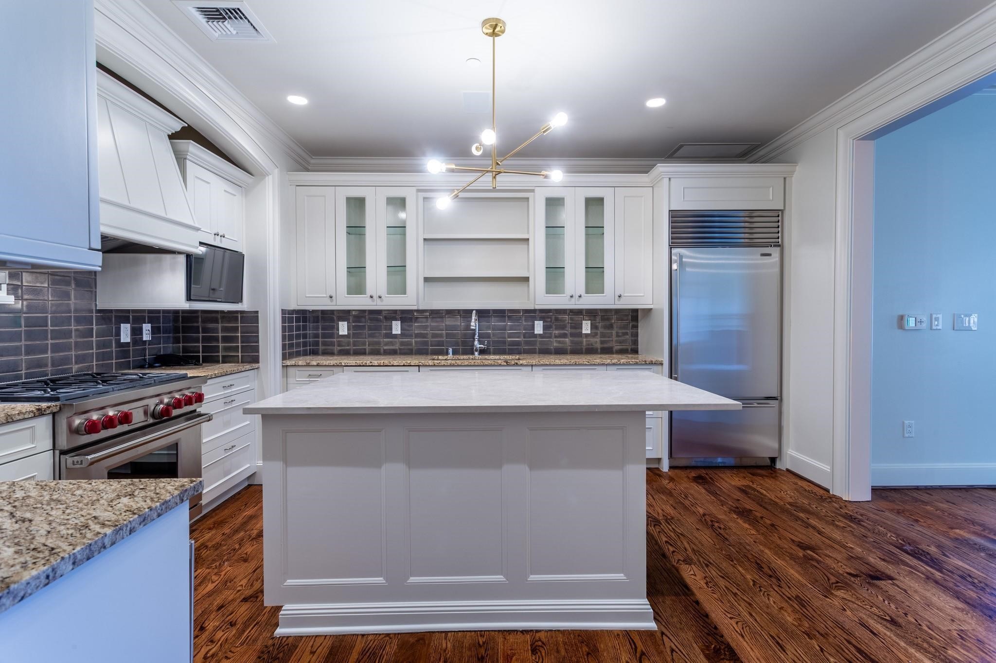 Kitchen with a large white island, stainless steel appliances, dark tile backsplash, and hardwood floors.