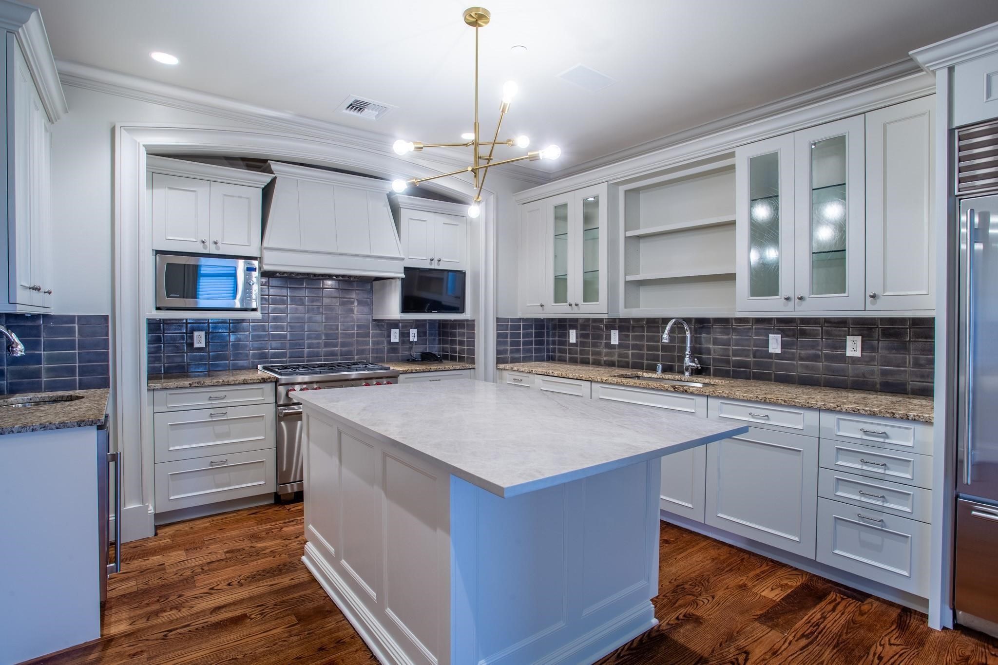 Kitchen with a large white island, dark tile backsplash, and warm wood floors.