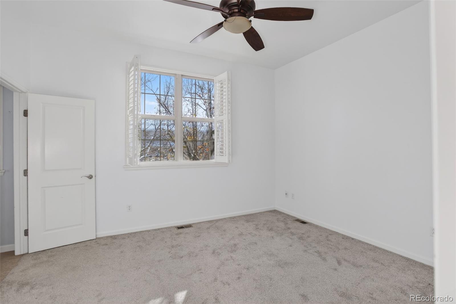 Empty bedroom with gray carpet, ceiling fan, and shuttered window.