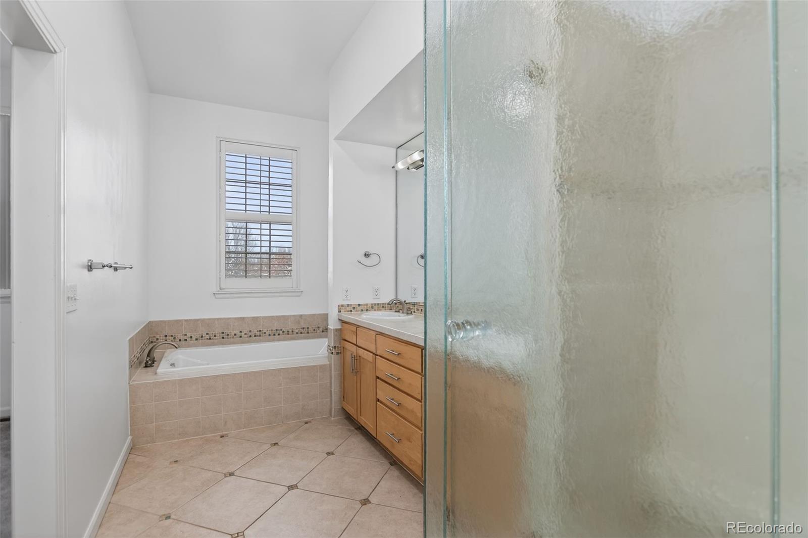 Bathroom with a soaking tub beneath a shuttered window and a wood vanity beside a glass shower.