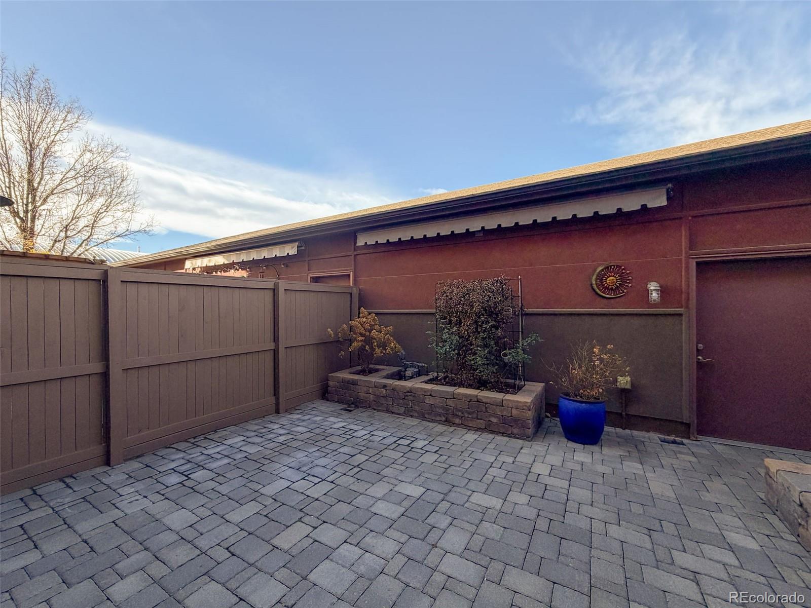Paved patio with a privacy fence, raised planter, and blue potted plant beside the home.