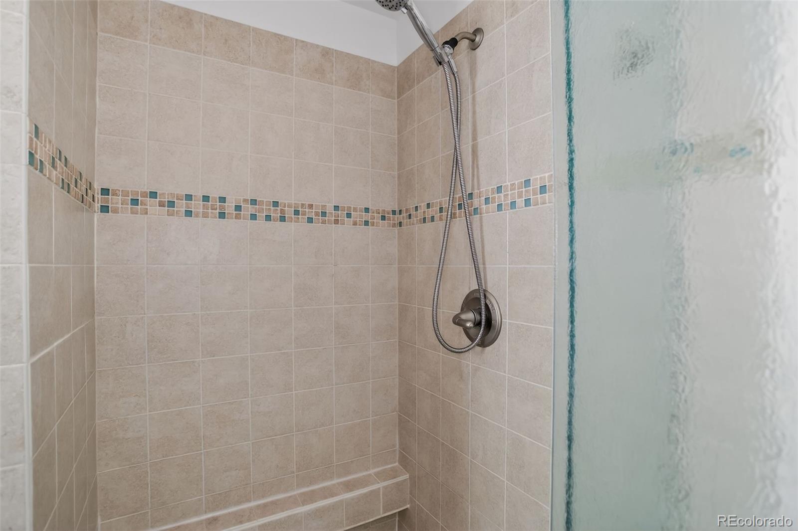 Walk-in shower with beige tile, a mosaic accent band, handheld showerhead, and frosted glass partition.