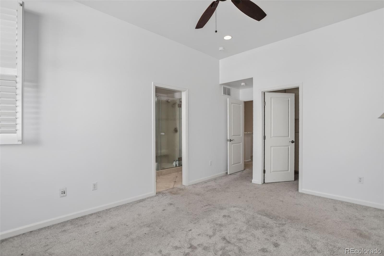 Empty carpeted bedroom with a ceiling fan, plantation shutters, and an open doorway to a glass shower.