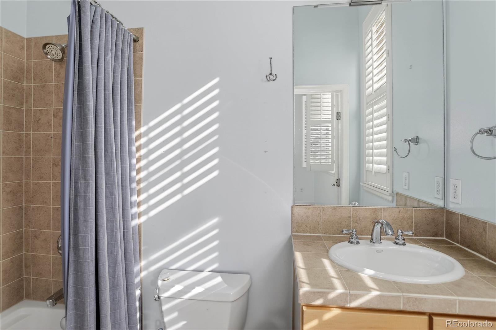 Bathroom with tan tile, gray shower curtain, and a vanity sink lit by sunlight through shutters.