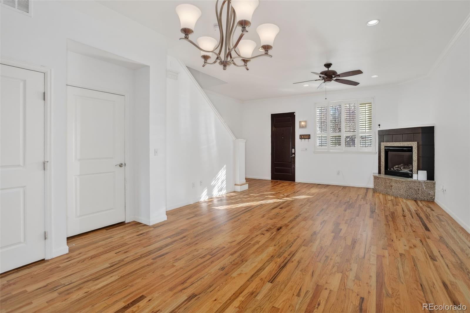 Vacant living room with hardwood floors, a fireplace, plantation shutters, and a ceiling fan.