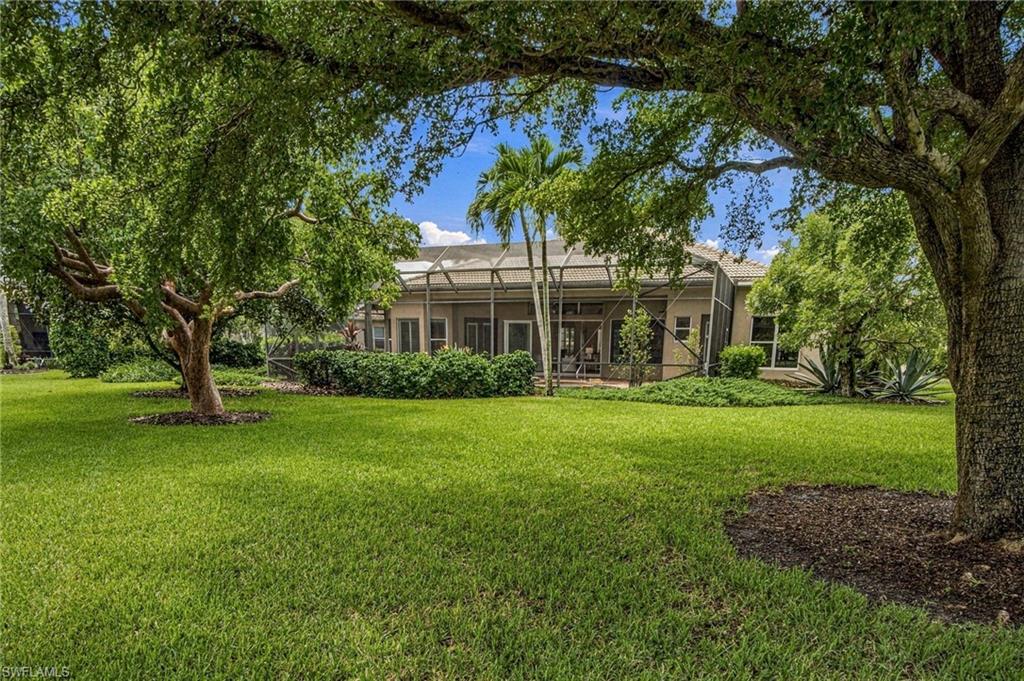 Rear of a single-story home behind a broad lawn, framed by mature trees and tropical landscaping.