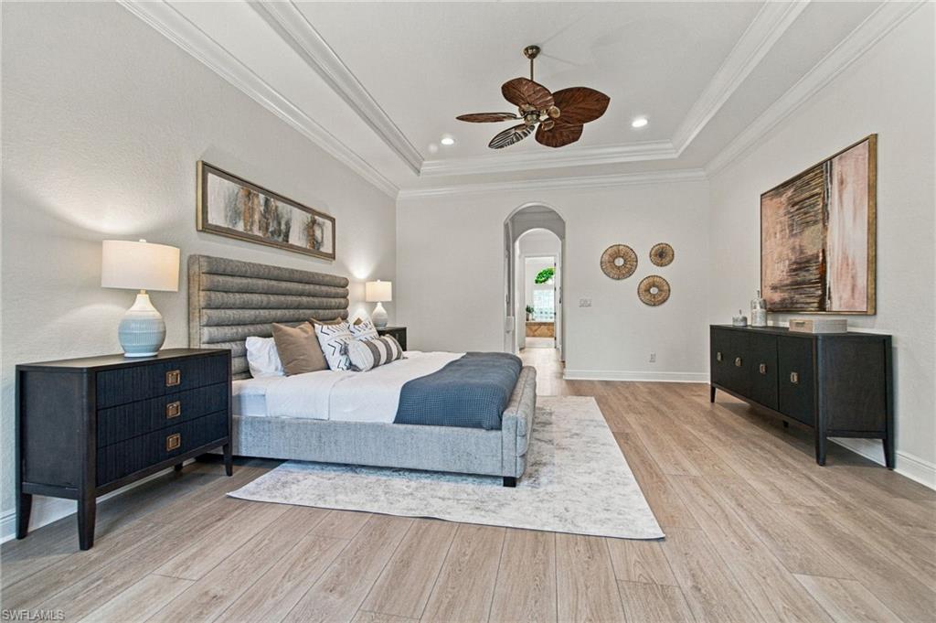 Furnished bedroom with a gray upholstered bed, tray ceiling, dark wood furniture, and arched doorway.