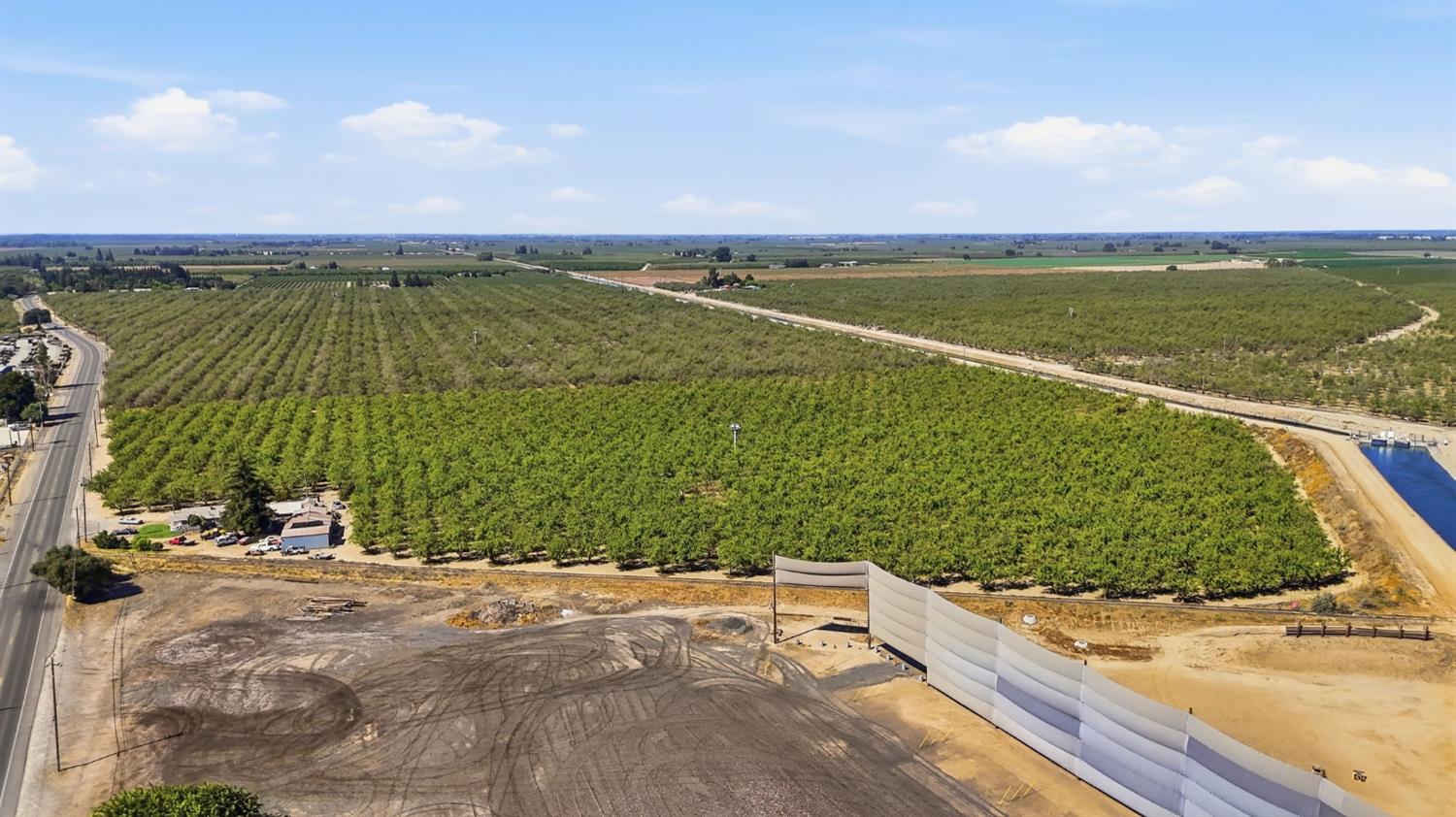 Aerial view of a green orchard bordered by a road and irrigation canal, with graded dirt in the foreground.