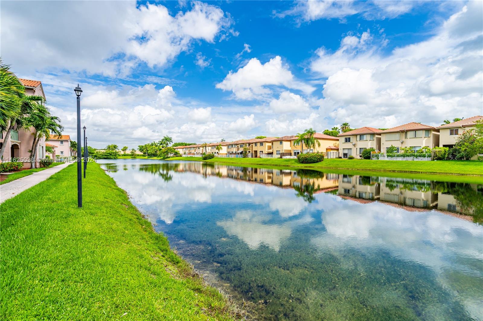 Residential homes line a calm lake, their reflections visible beside a grassy bank and palm trees.