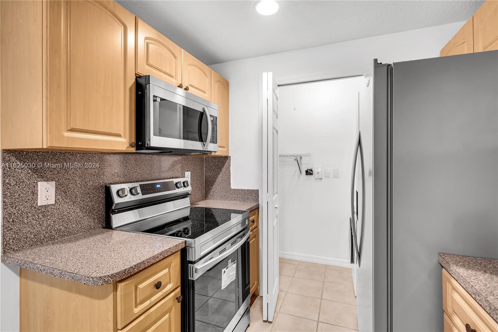Kitchen with light wood cabinets, speckled countertops, stainless steel appliances, and an open doorway.