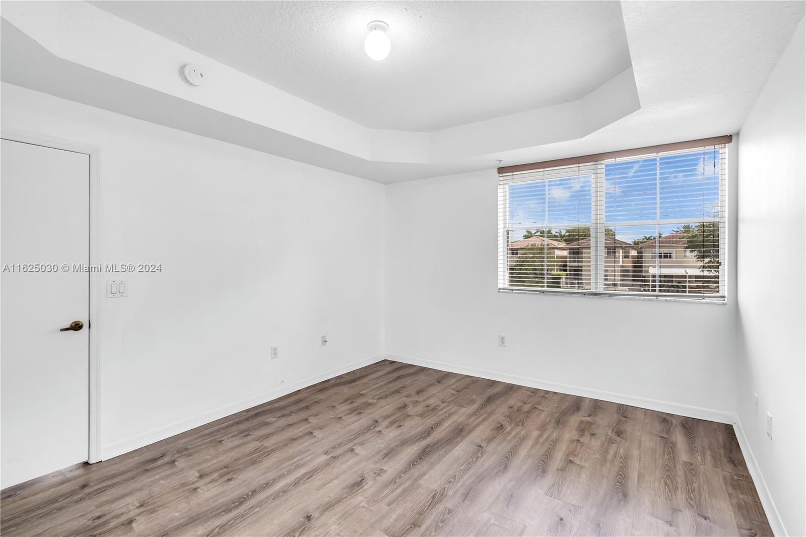 Empty room with wood-look flooring, a tray ceiling, and a large window overlooking nearby homes.