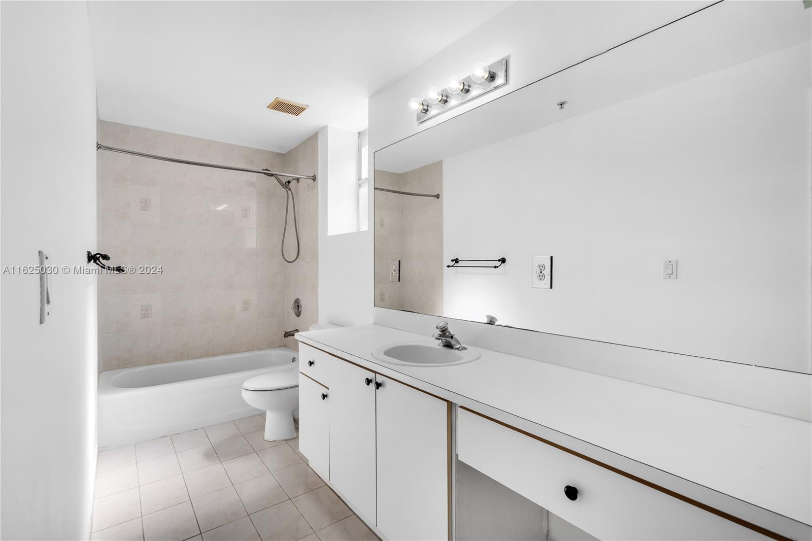 Bathroom with a tiled tub and shower, toilet, and long white vanity beneath a large mirror.