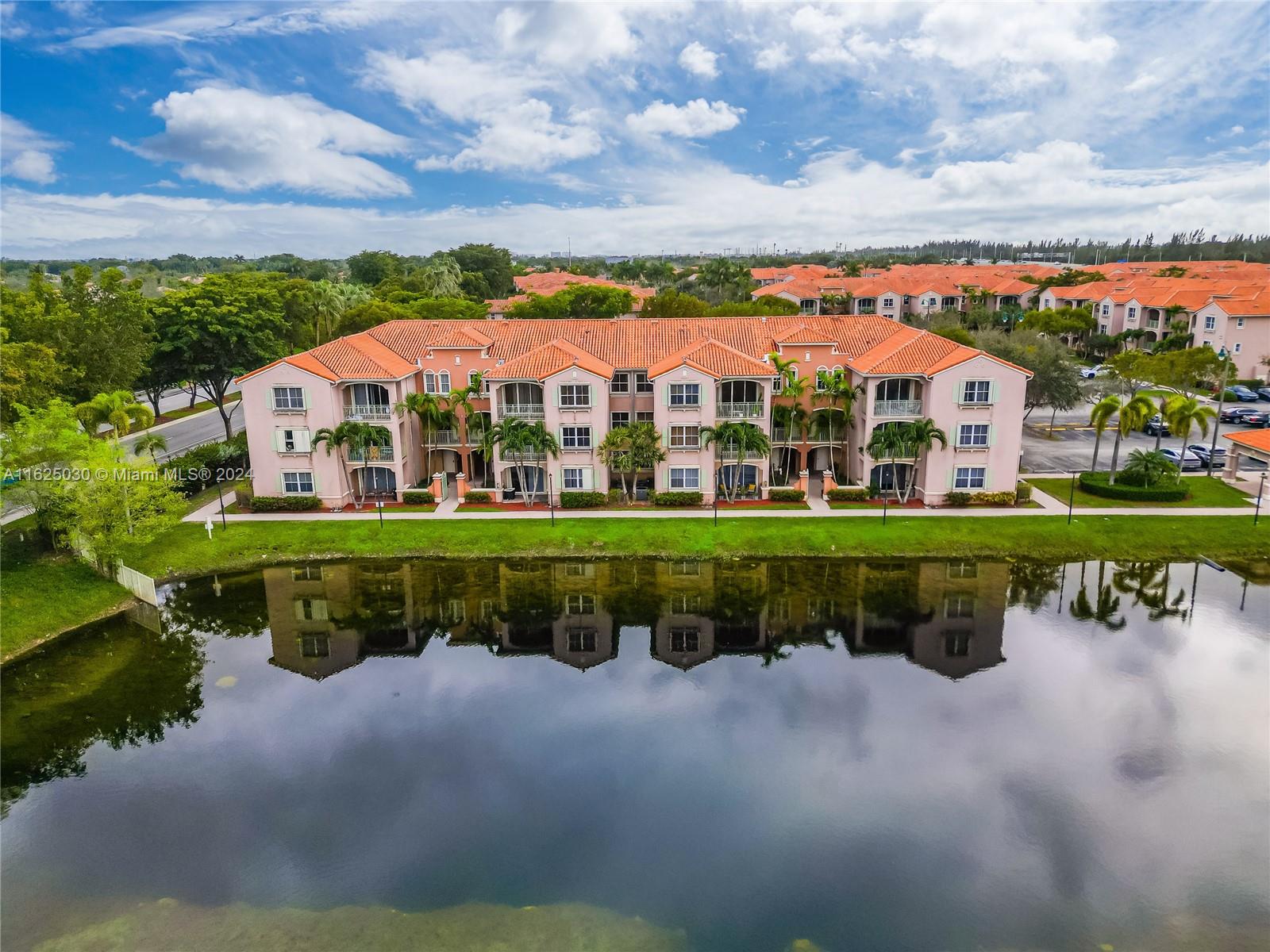 Pink condominium building with terracotta roofs reflected in a pond, viewed from above.