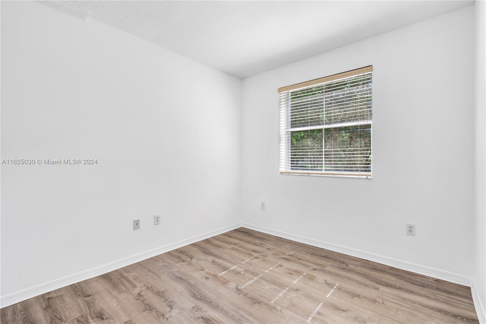 Vacant room with wood-look flooring and a window with blinds overlooking greenery.