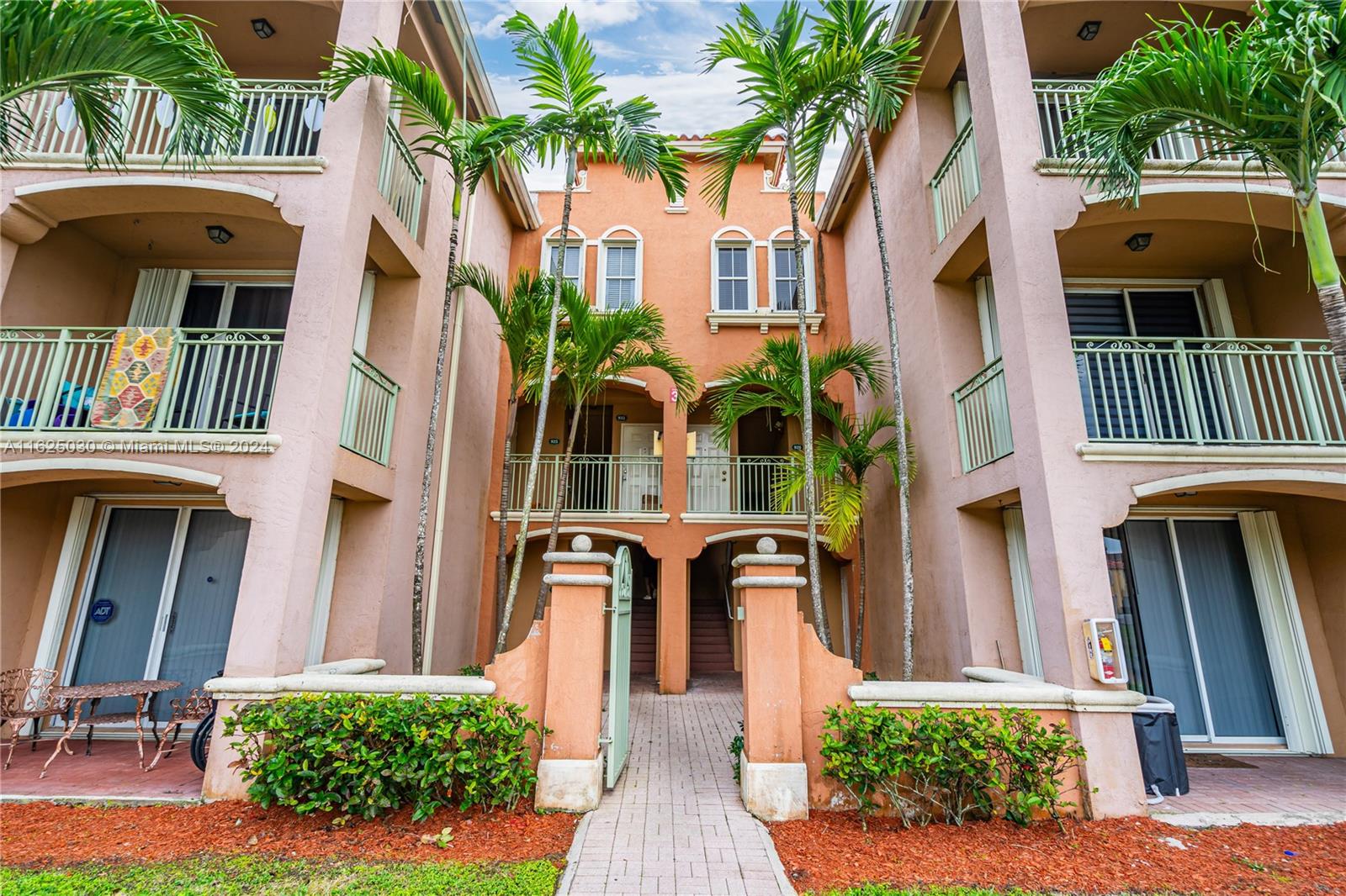 Peach condominium building with palm-lined entrance, central walkway, balconies, and ground-floor patios.
