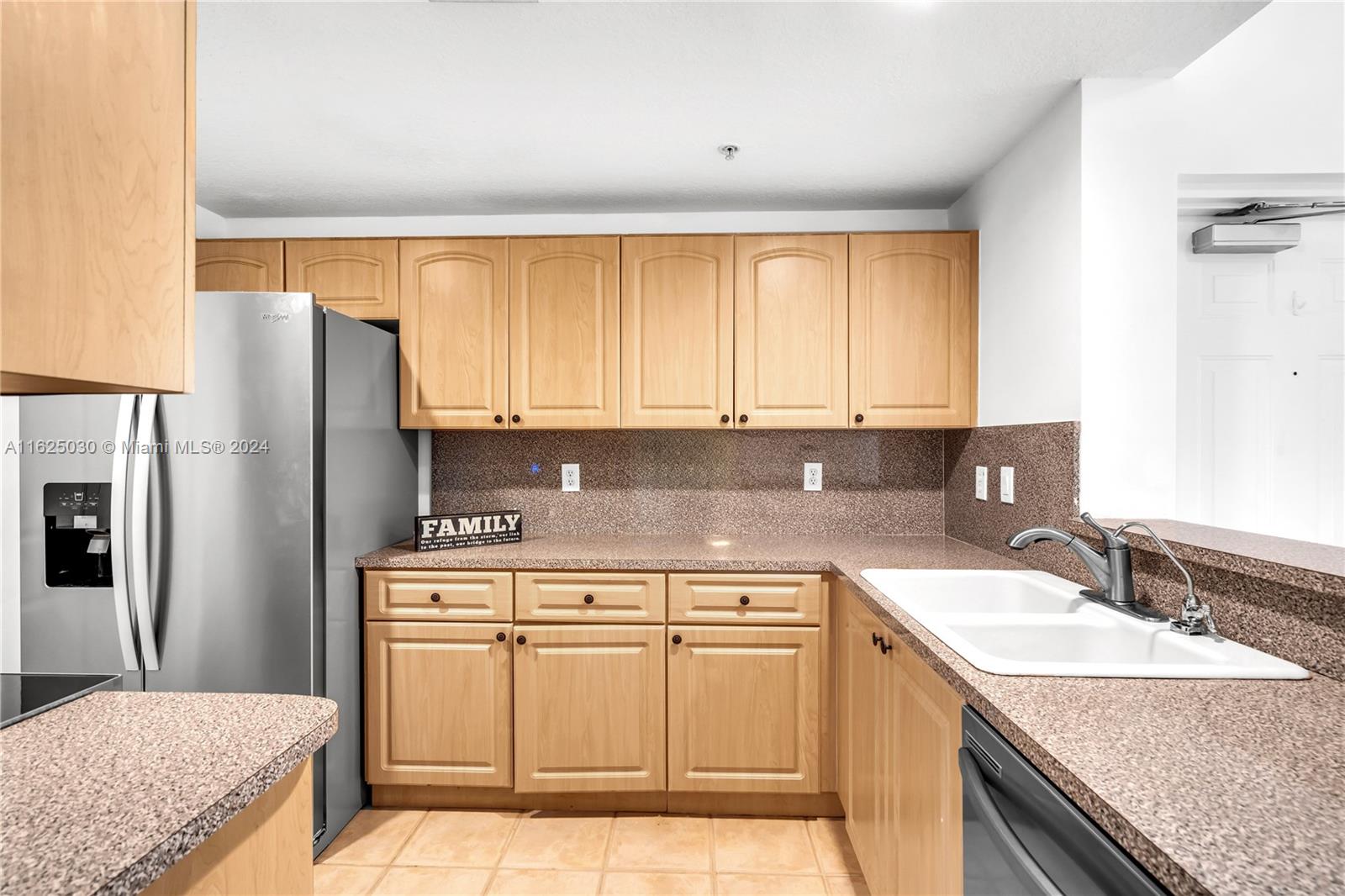 Kitchen with light wood cabinetry, speckled countertops, stainless refrigerator, and white double-basin sink.