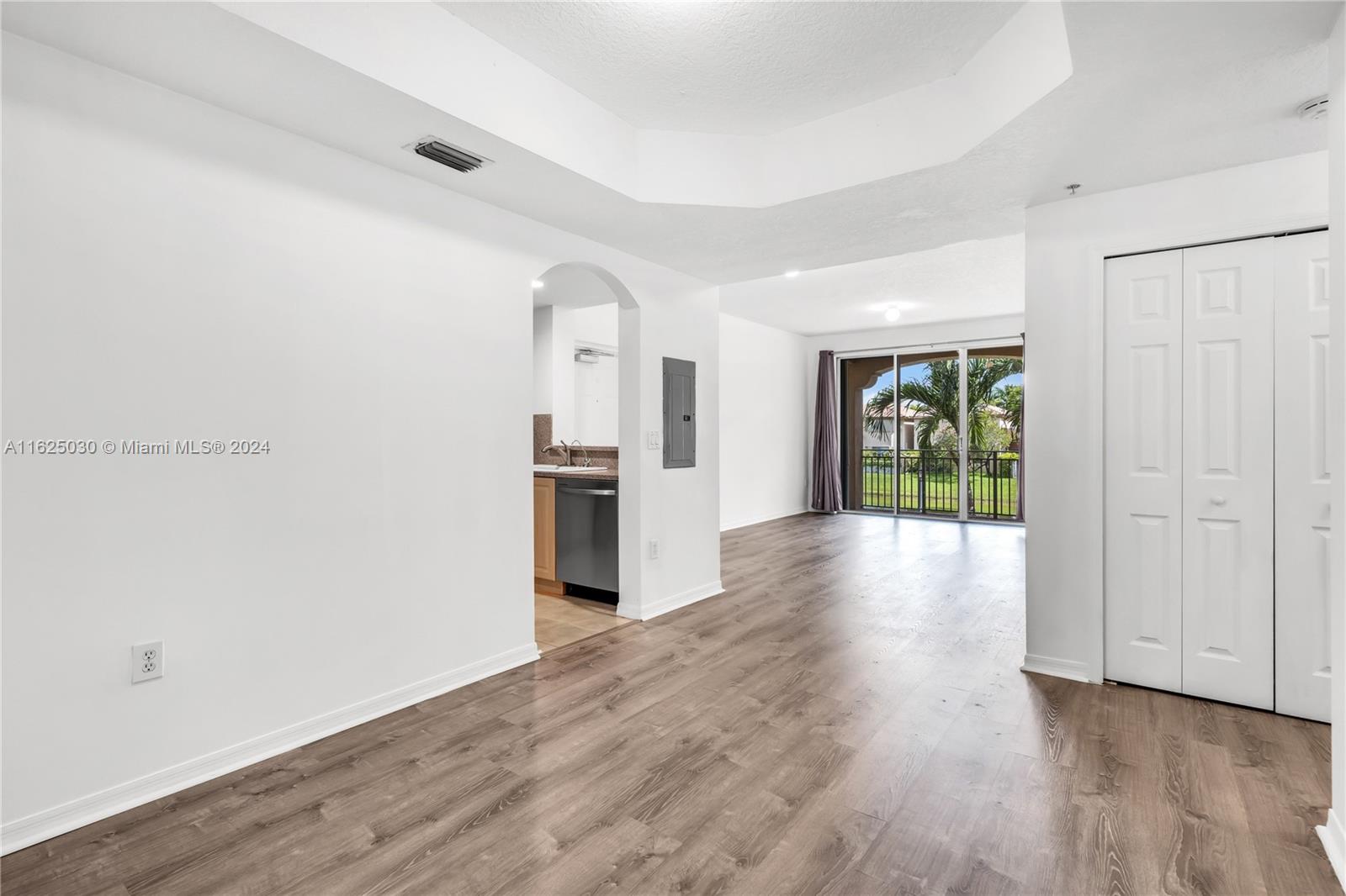 Vacant living area with wood-look floors, an arched kitchen opening, and sliding doors overlooking a lawn and palms.