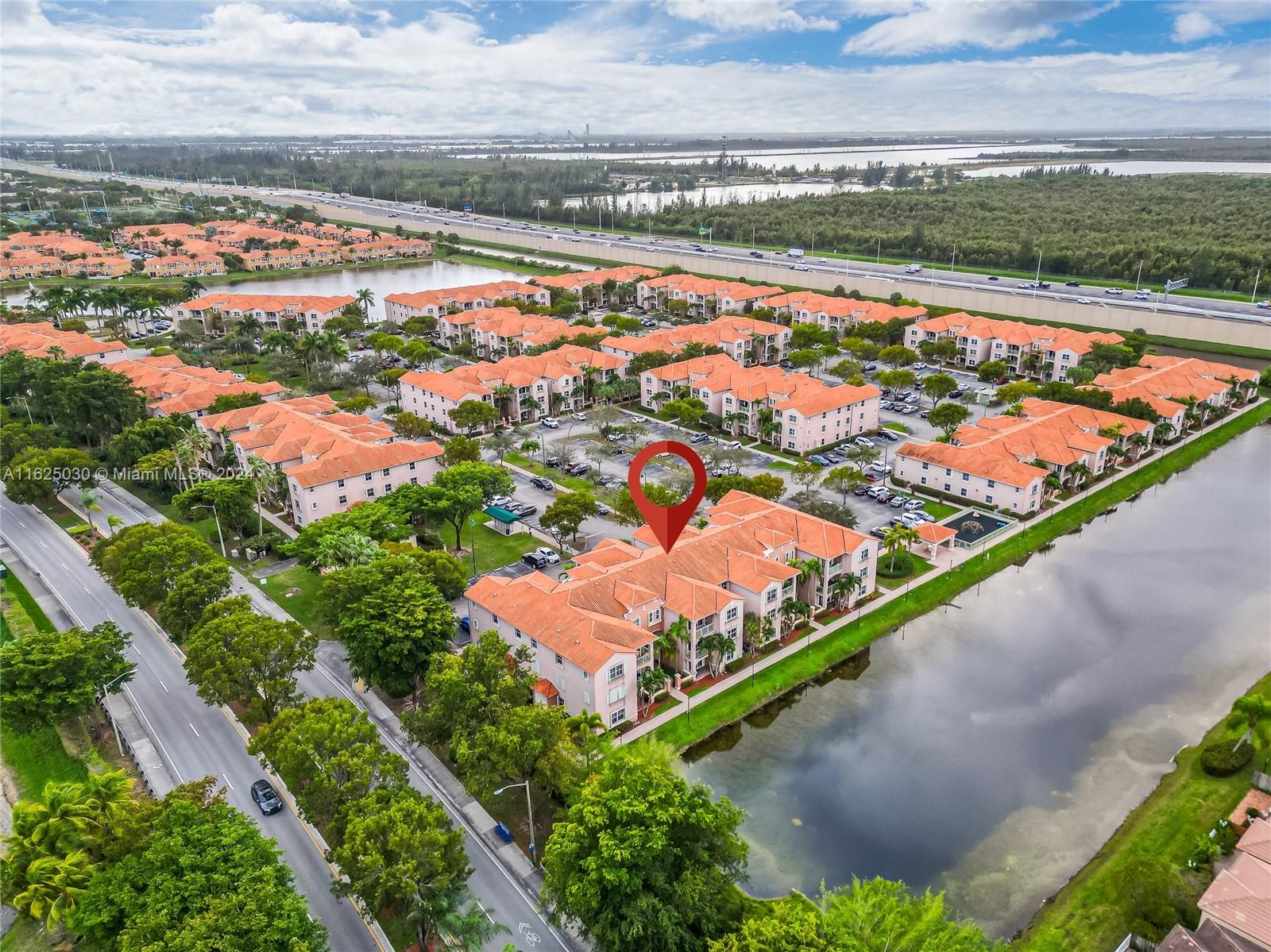 Aerial view of a condominium community with orange tile roofs, a pond, and a red location pin marking a building.