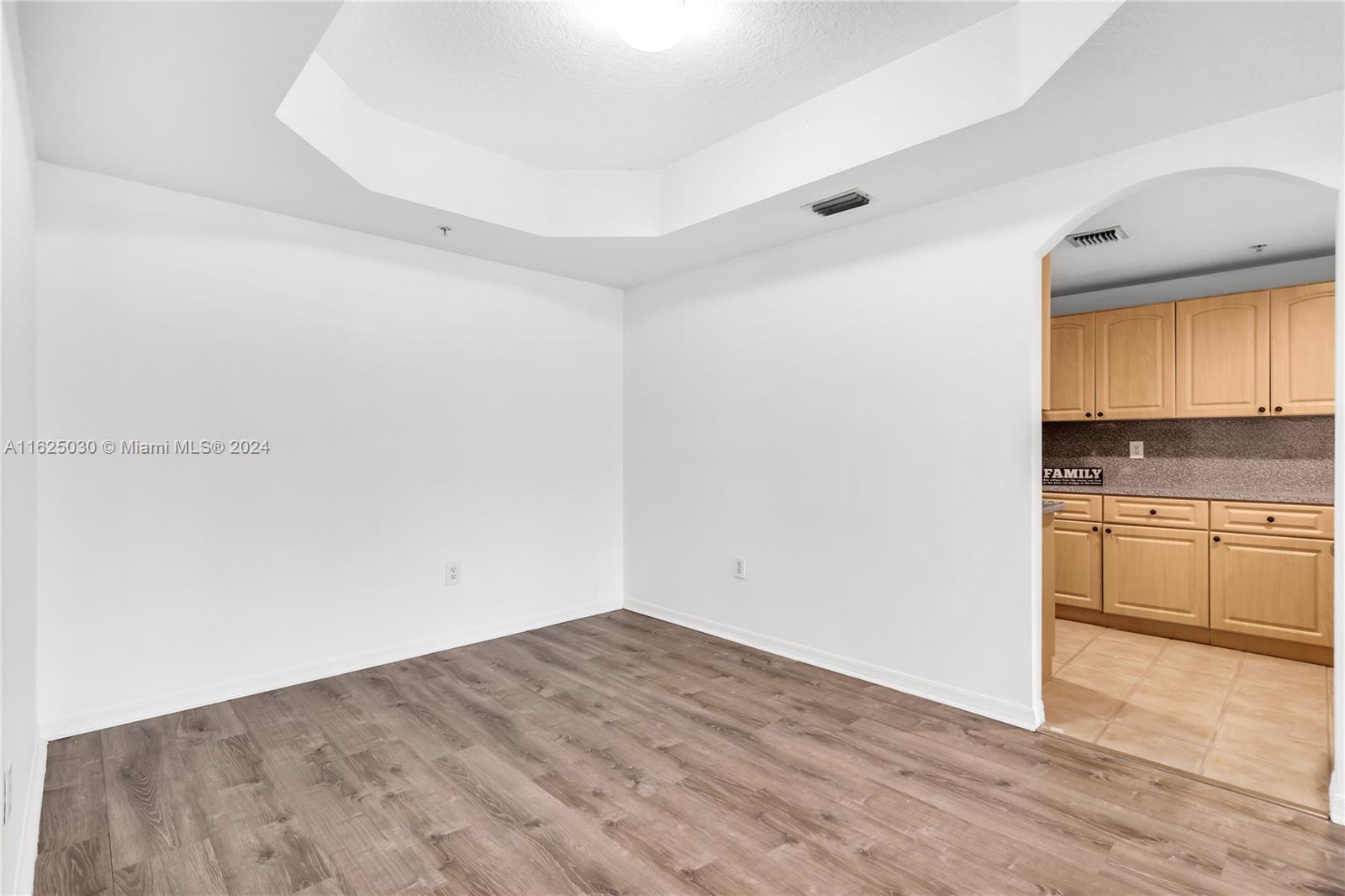 Vacant room with wood-look flooring, a tray ceiling, and an arched opening to the kitchen.