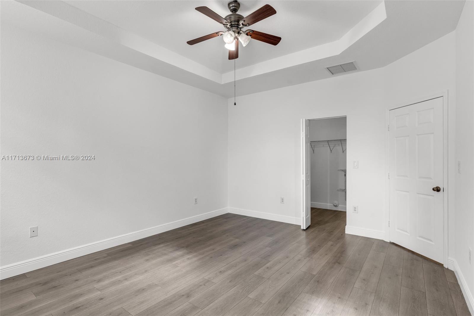 Vacant bedroom with gray plank flooring, tray ceiling, ceiling fan, and open closet.