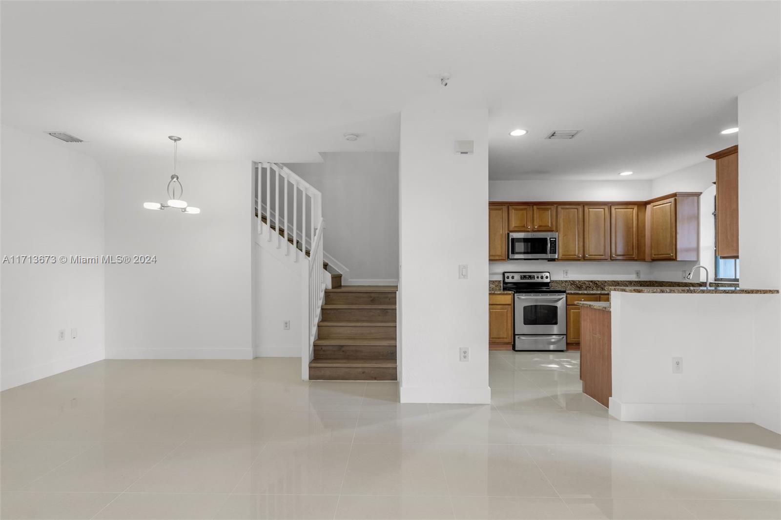 Open living area with tiled floors, staircase, and adjoining kitchen with wood cabinets and stainless-steel appliances.