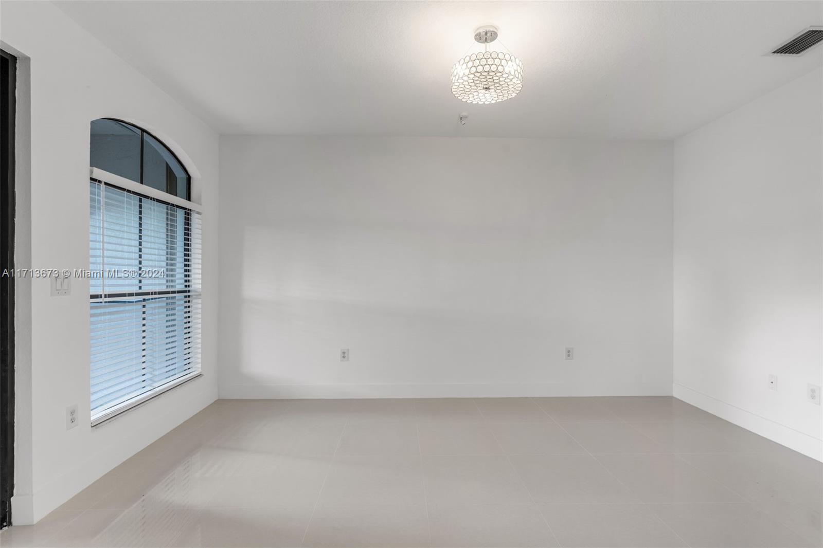 Vacant bright room with glossy tile floors, an arched window with blinds, and a decorative ceiling light.