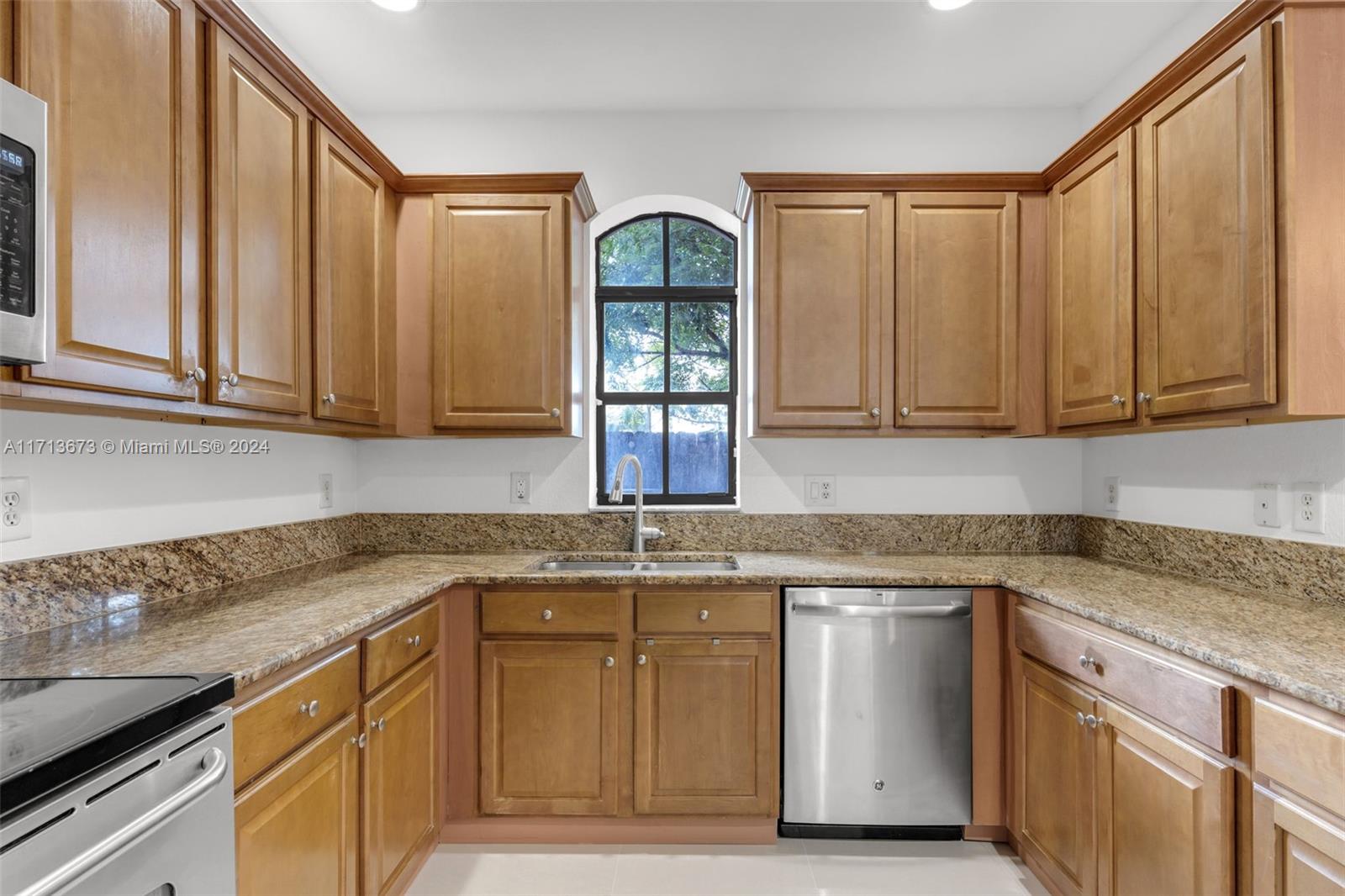 Kitchen with wood cabinets, granite counters, stainless-steel dishwasher, and an arched window above the sink.