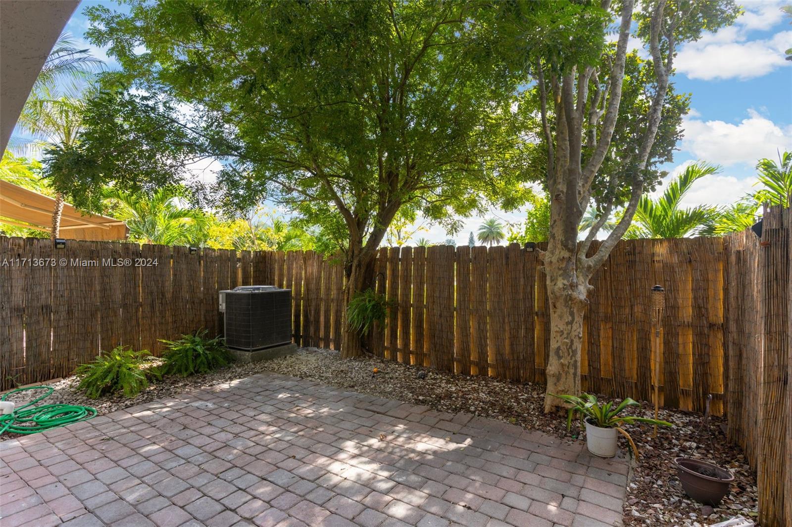 Shaded paved patio enclosed by a bamboo fence, with mature trees and potted plants.