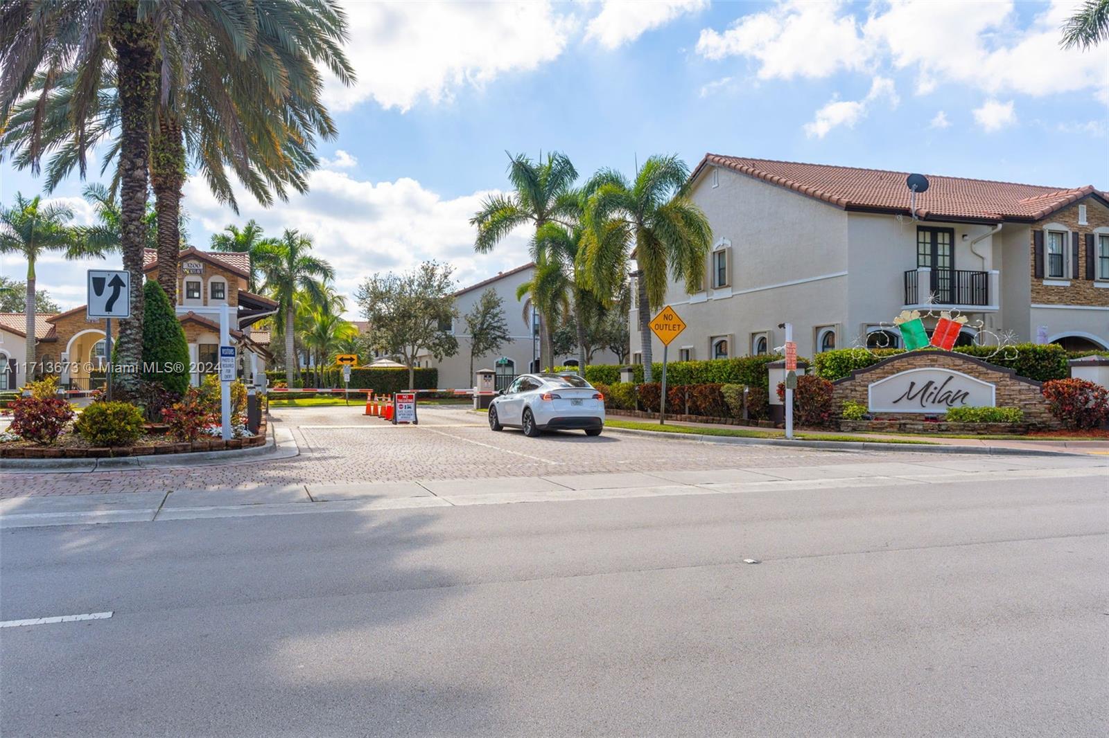 Gated Milan community entrance with palm trees, a stone sign, and Mediterranean-style residences.
