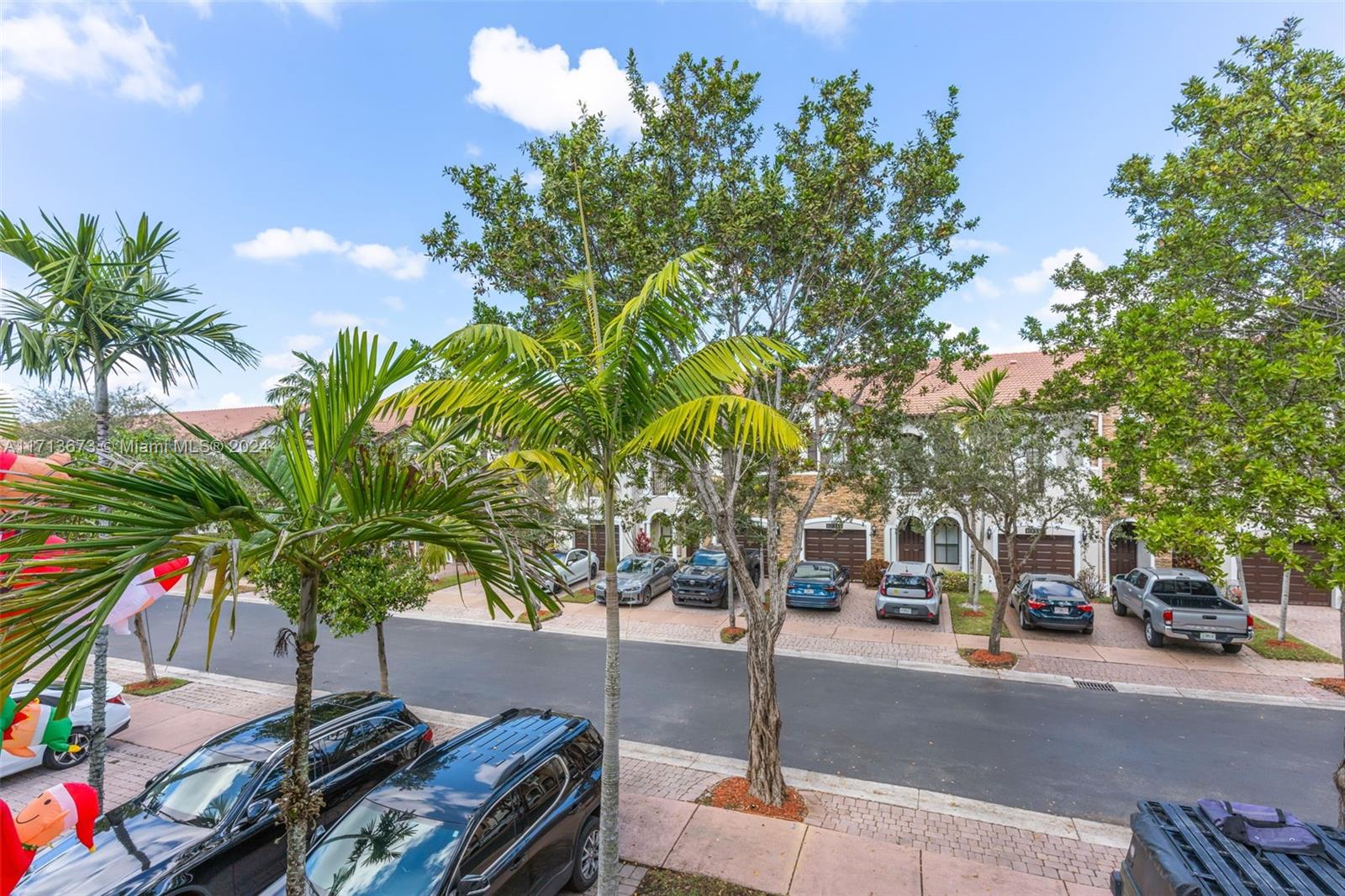Attached homes with garages line a tree-shaded street with parked cars and tropical landscaping.