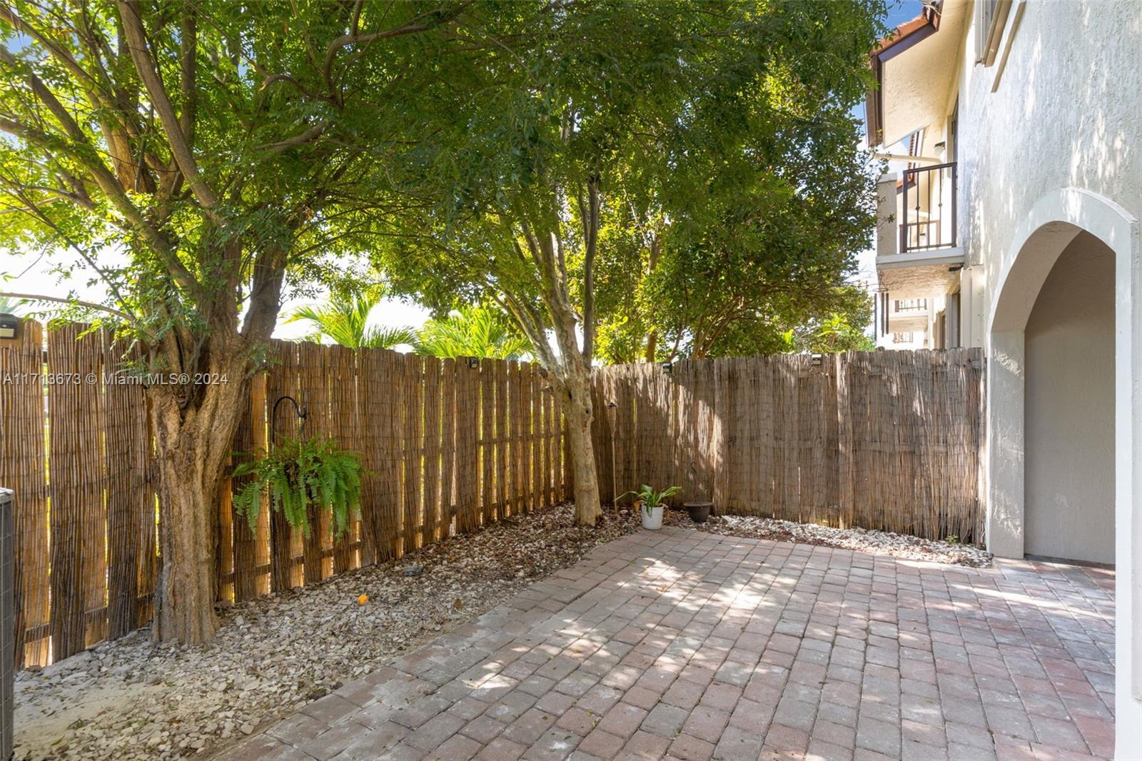 Private brick-paved patio with mature shade trees, reed fencing, and an arched passage beside the home.