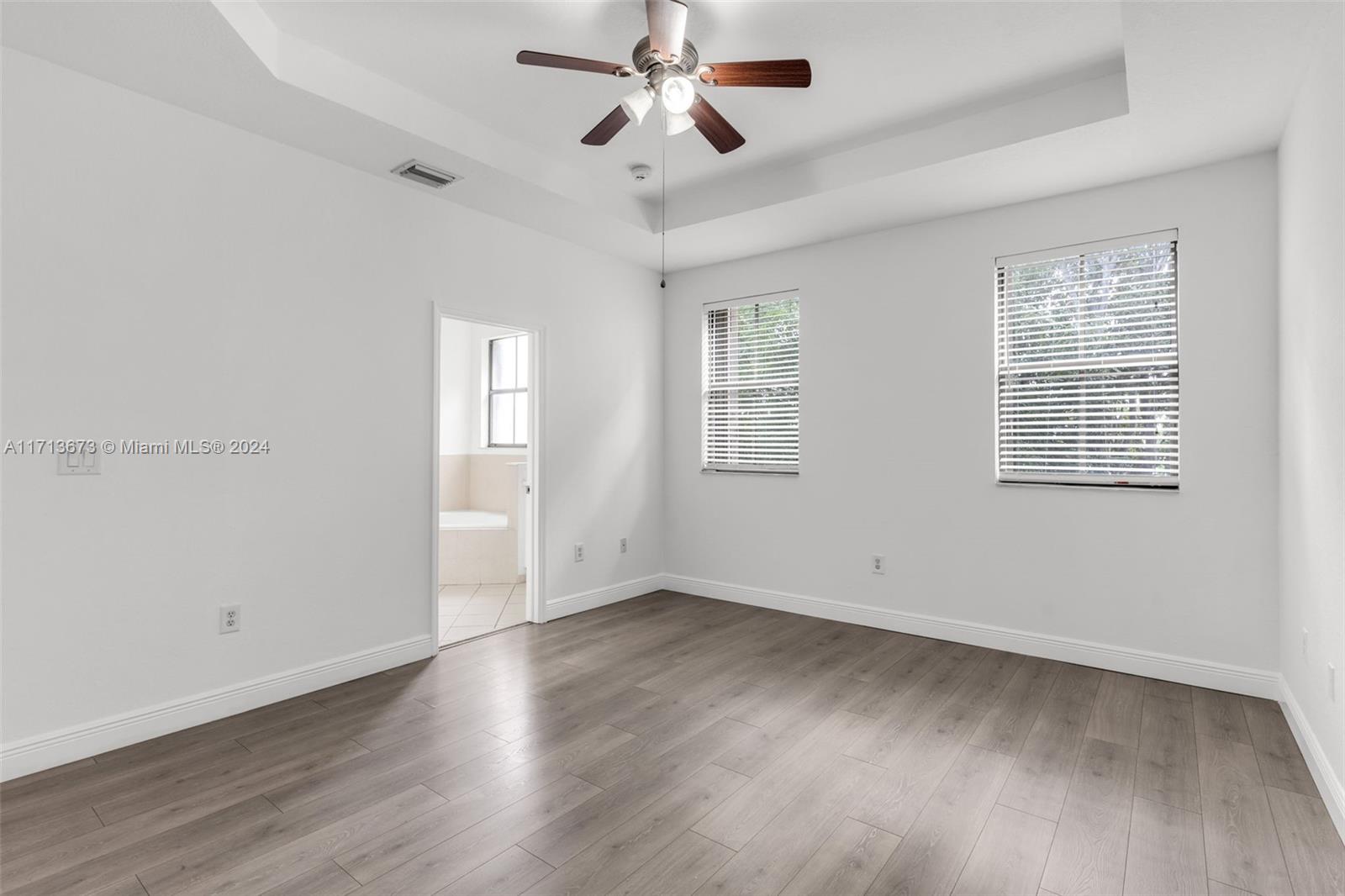 Vacant bedroom with gray plank flooring, a ceiling fan, and windows with blinds; bathroom visible through doorway.