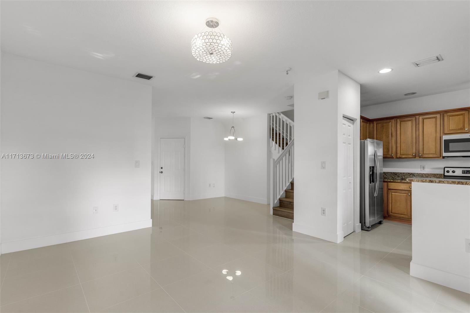 Vacant open-plan living and dining area with glossy tile floors, wood kitchen cabinets, and a staircase.