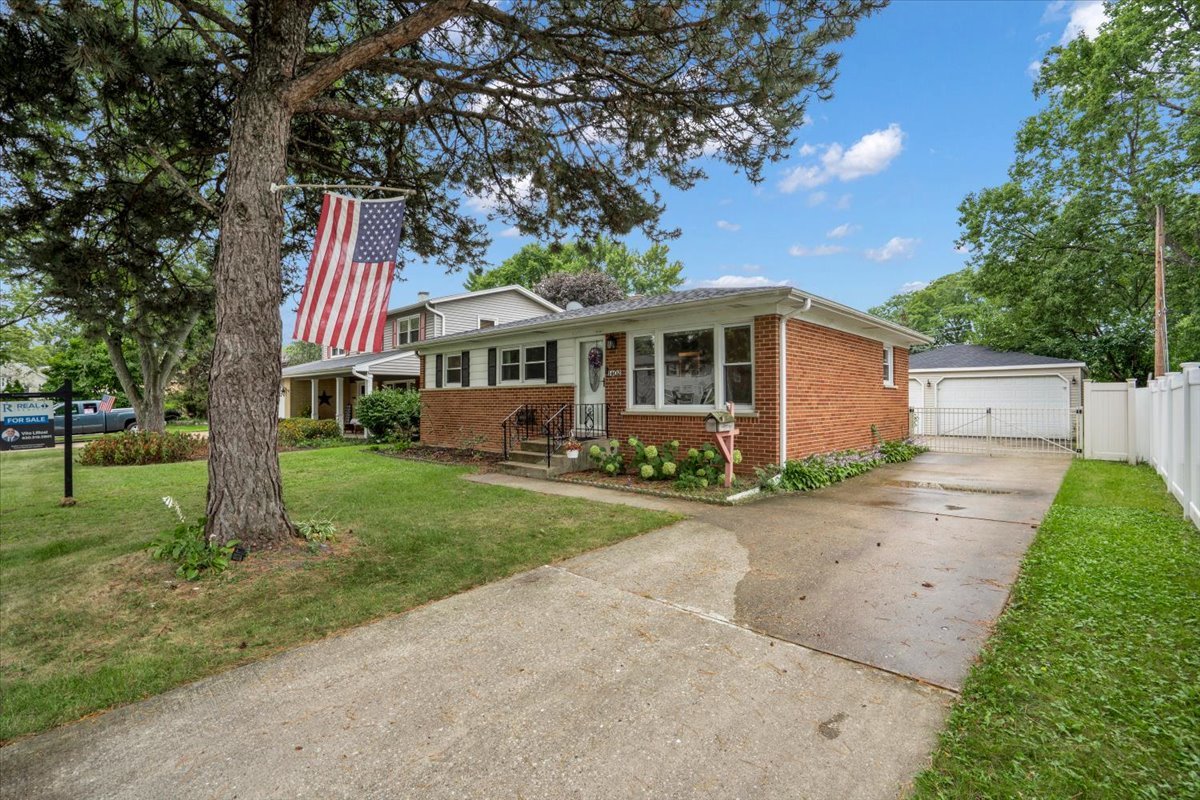 Brick ranch house with a large American flag, mature trees, and a driveway to a detached garage.