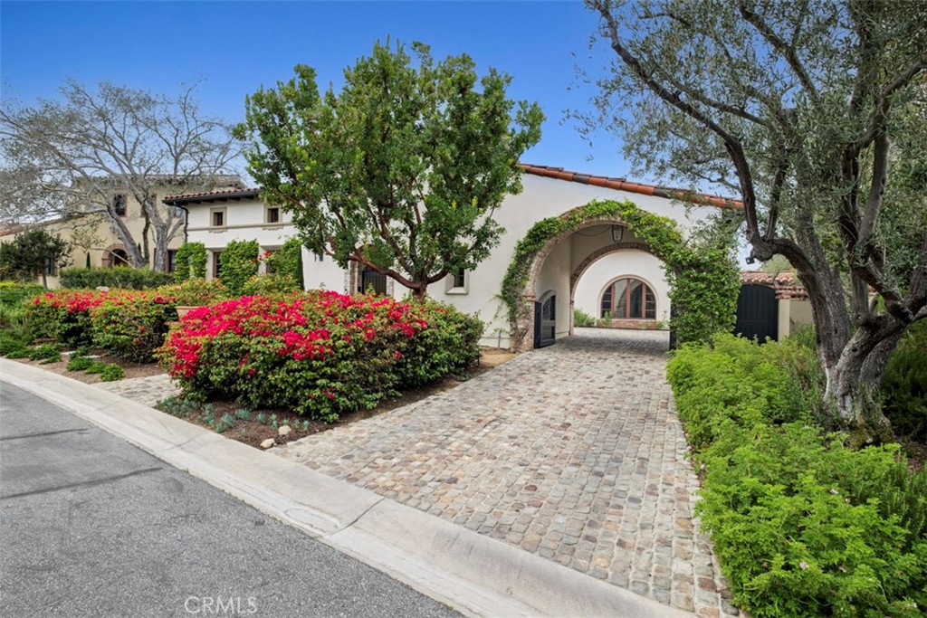 White stucco home with an arched entry, cobblestone driveway, and flowering shrubs.