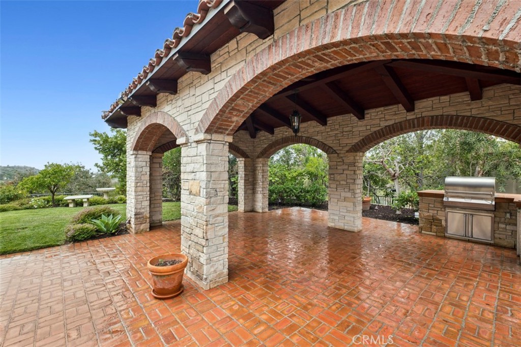 Covered brick patio with stone arches, exposed wood beams, and a built-in stainless steel grill.