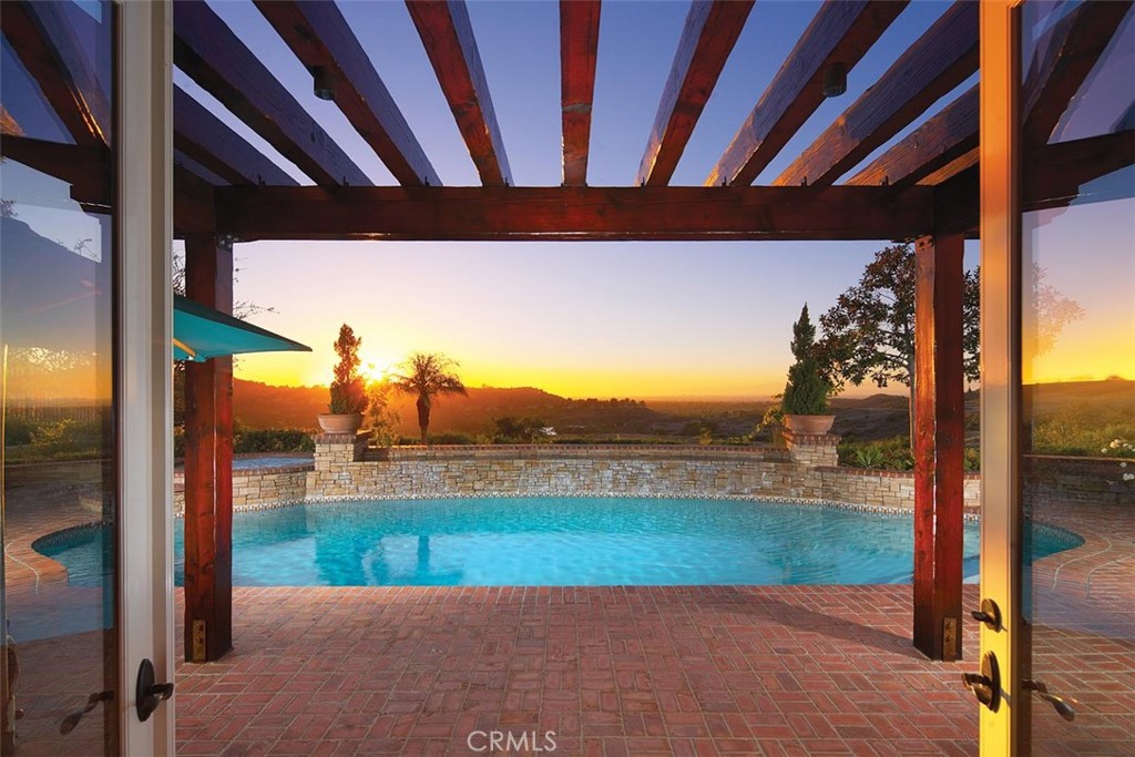 Swimming pool beneath a timber pergola, with a brick patio and sunset view over hills.