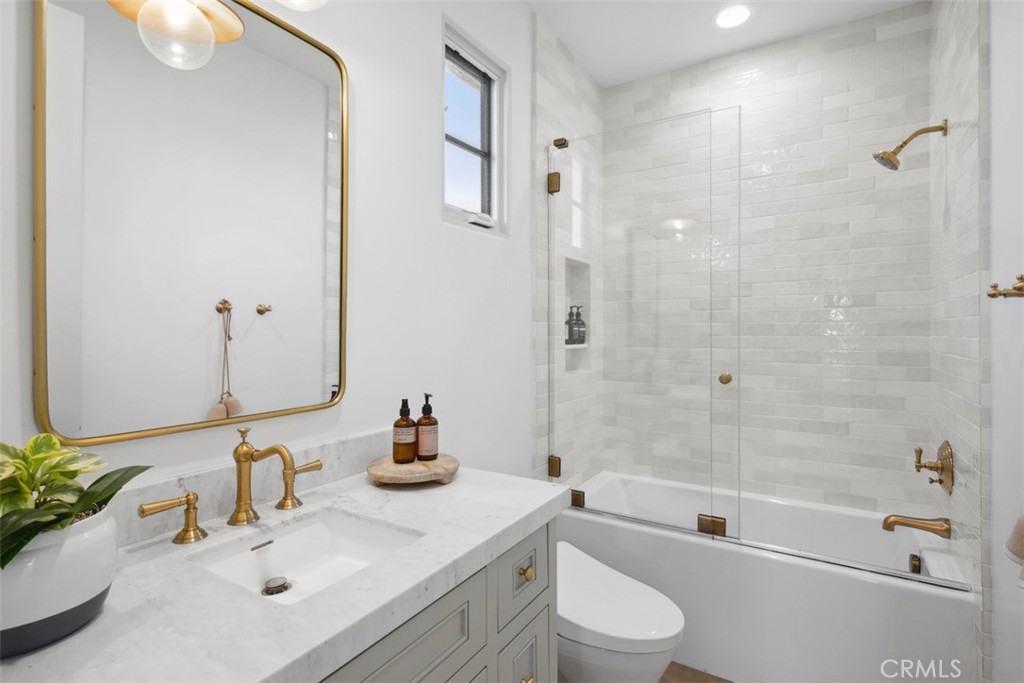 Bright bathroom with a marble vanity and brass fixtures beside a glass-enclosed, white-tiled bathtub.