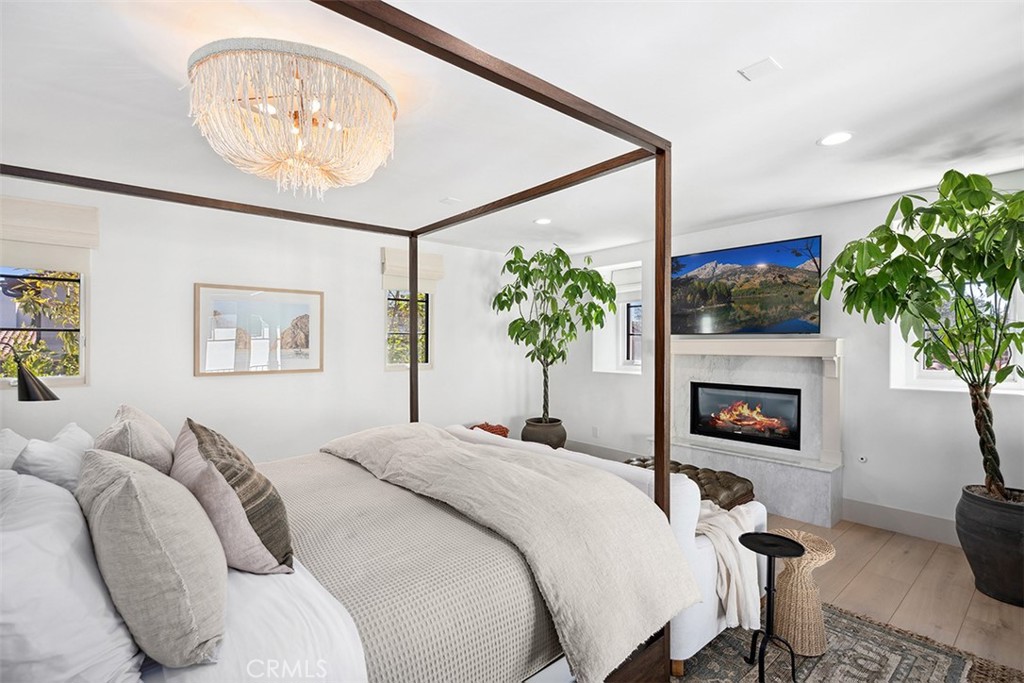 Bright bedroom with a dark wood canopy bed, chandelier, and fireplace beneath a wall-mounted TV.