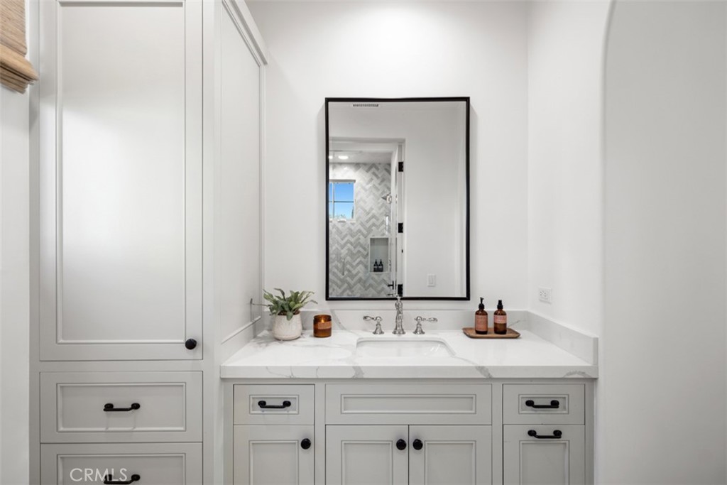 Bathroom vanity with white paneled cabinets, a marble-look counter, and a mirror reflecting a herringbone-tile shower.