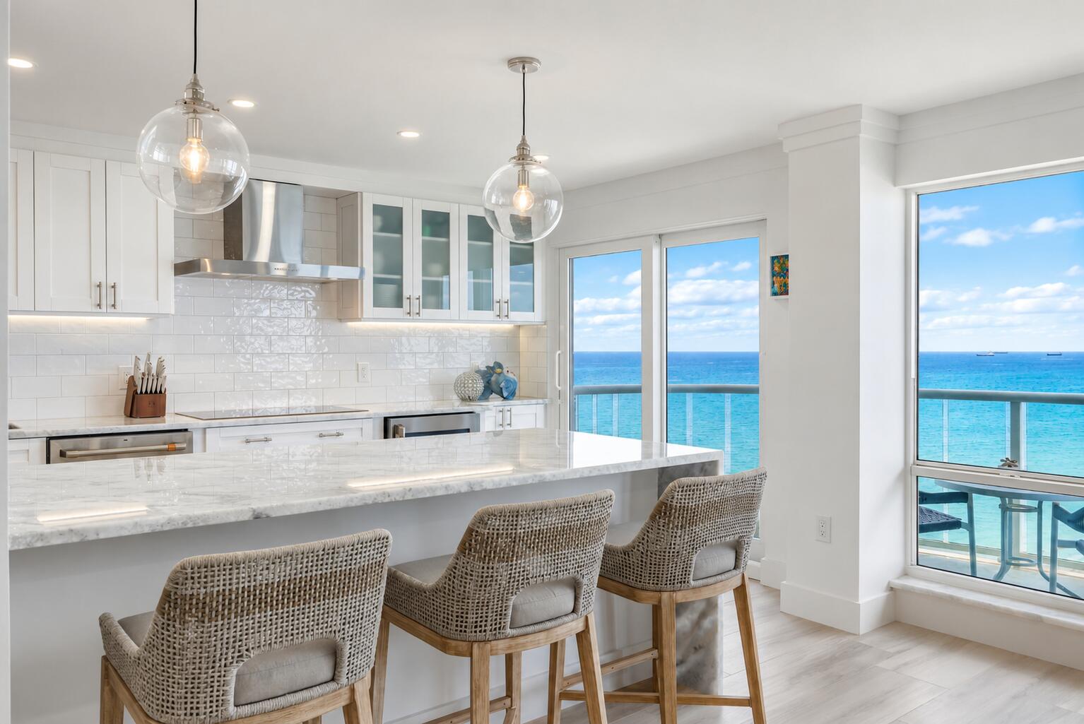 Modern white kitchen with a marble island and ocean views through floor-to-ceiling windows.