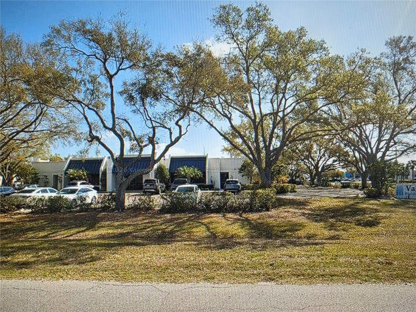 Modern commercial building behind parked cars, shaded by large trees.