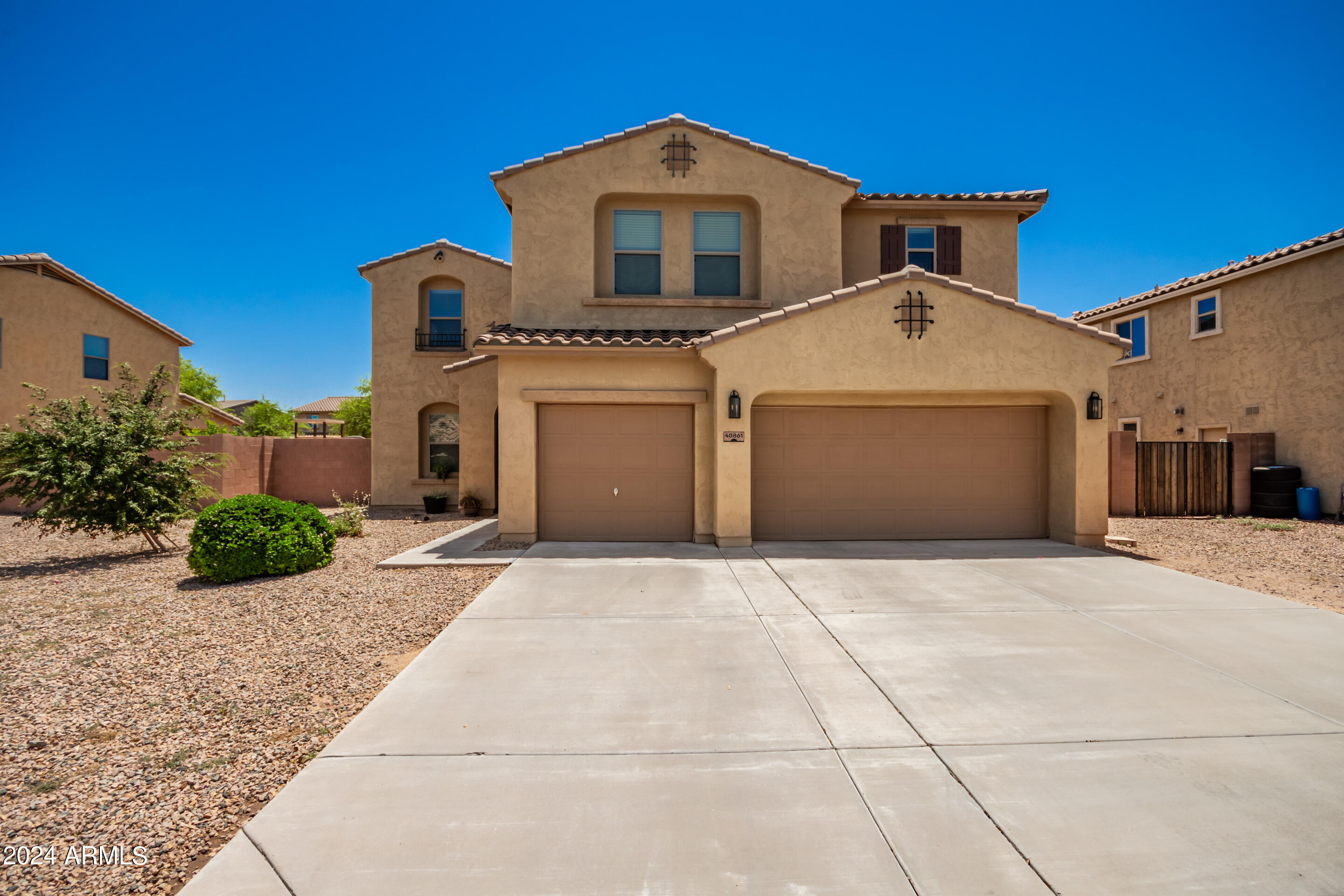 Two-story tan stucco home with a wide driveway, dual garage doors, and gravel landscaping.