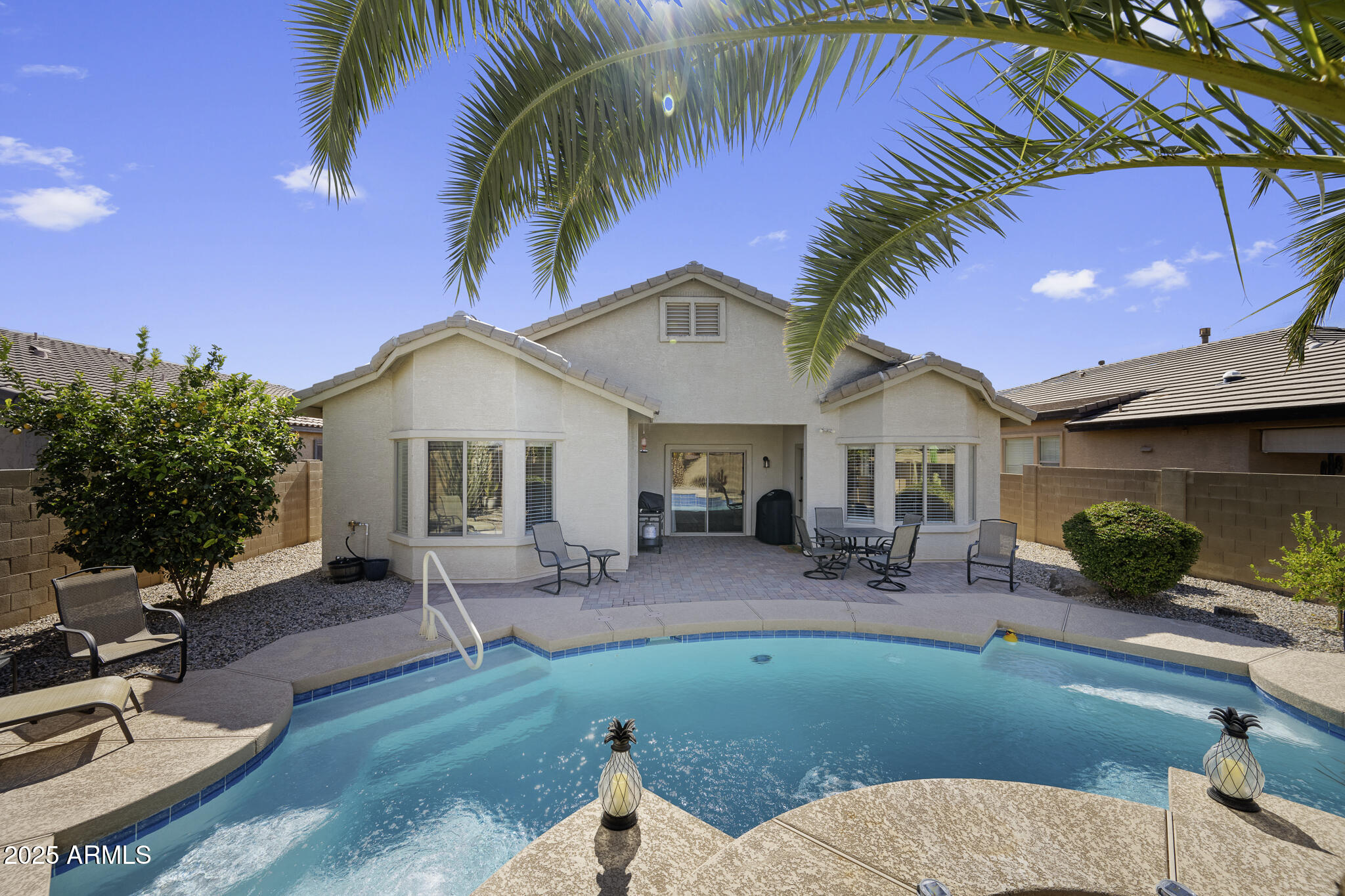 Home’s rear patio and seating overlook a turquoise swimming pool, framed by palm fronds.