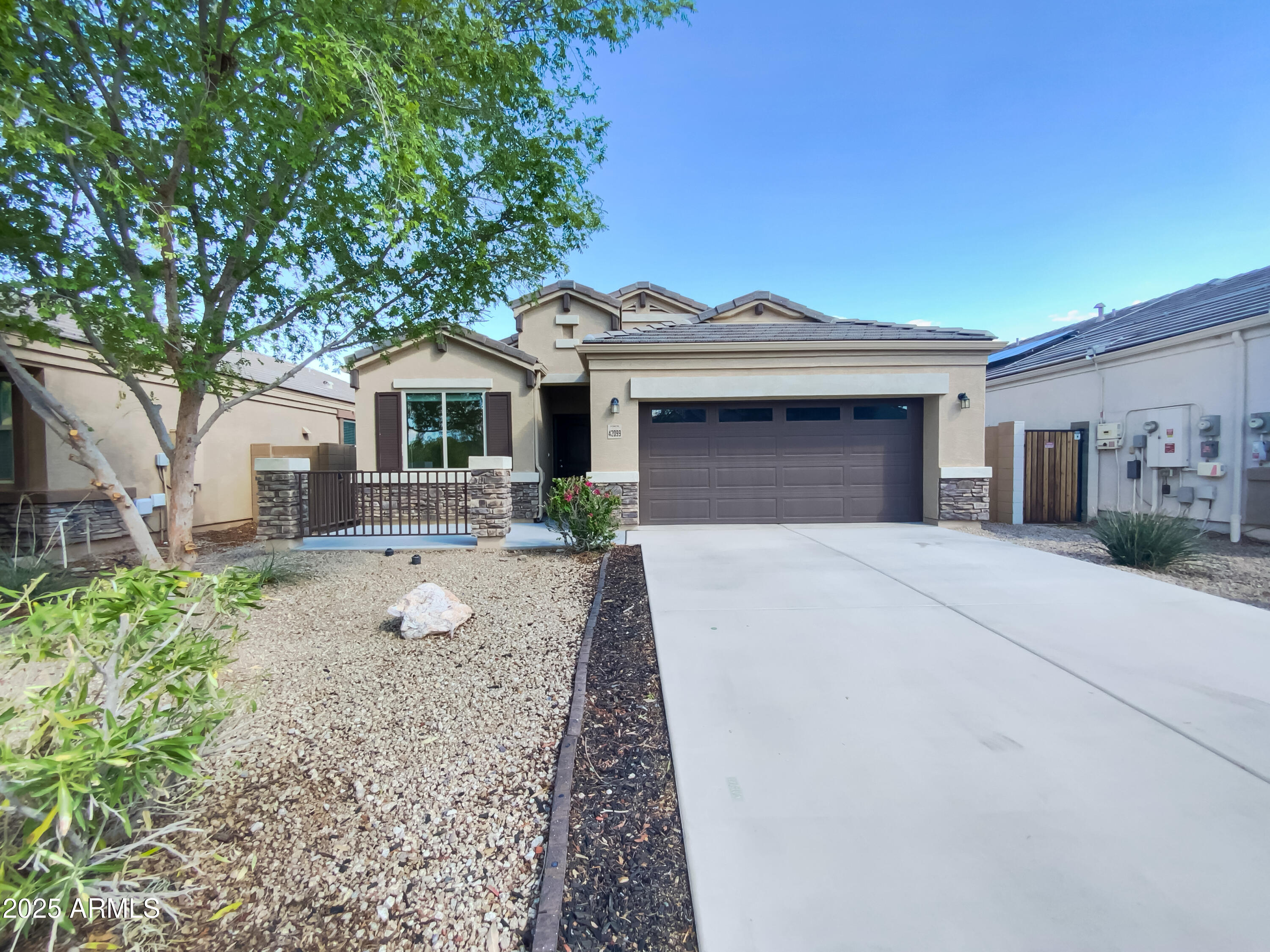 Beige stucco home with a dark double garage door, wide driveway, and gravel landscaping beneath a large tree.