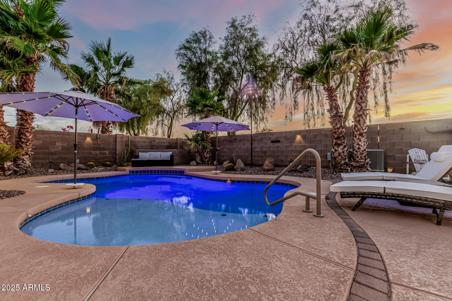 Backyard pool at dusk with purple umbrellas, palm trees, and white lounge chairs.
