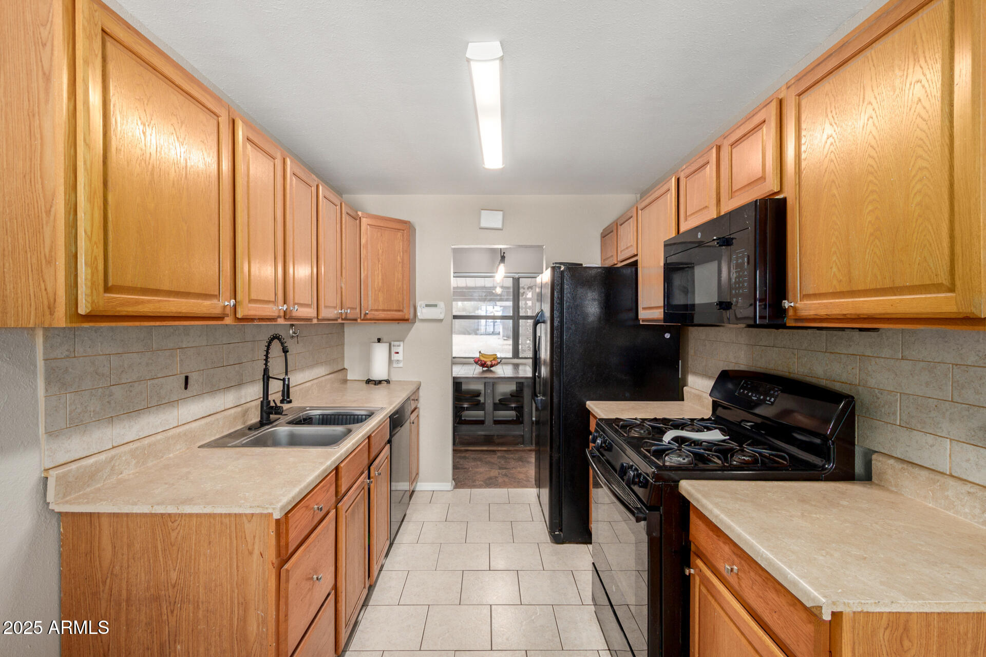 Galley kitchen with honey-colored cabinets, double sink, black appliances, and a window-side nook.