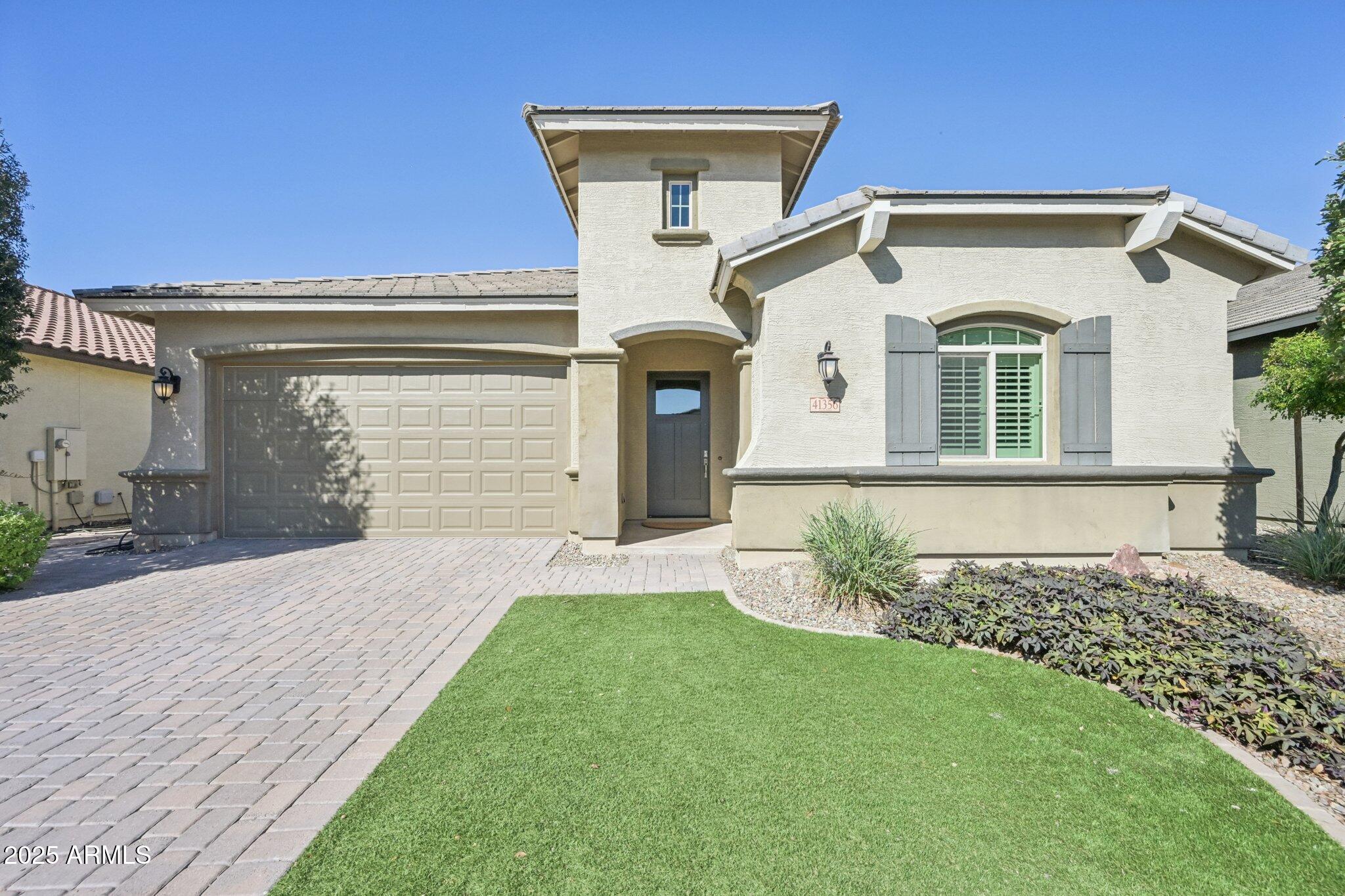 Cream stucco home with tower-like entry, gray shutters, paver driveway, and landscaped lawn.